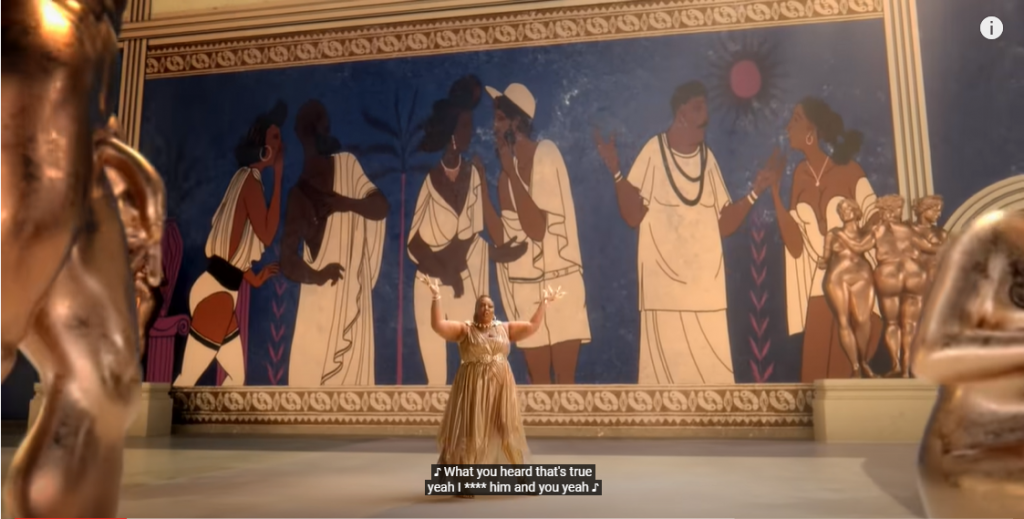 Lizzo in 0:20 of Rumors video, dressed in long gold dress, surrounded by large ancient Greek style murals or various people gossiping, with golden statue groups on edges of frame