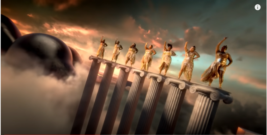 Lizzo and her dancers all dressed in flowing gold garments, dancing atop ionic columns with a mountain-sized butt far in the background
